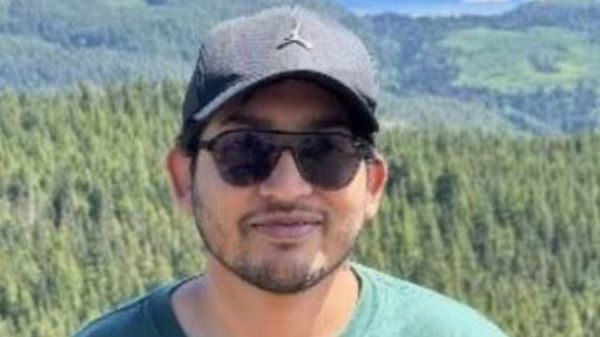 Man wearing a dark cap and sunglasses outdoors with a forested mountainside behind him