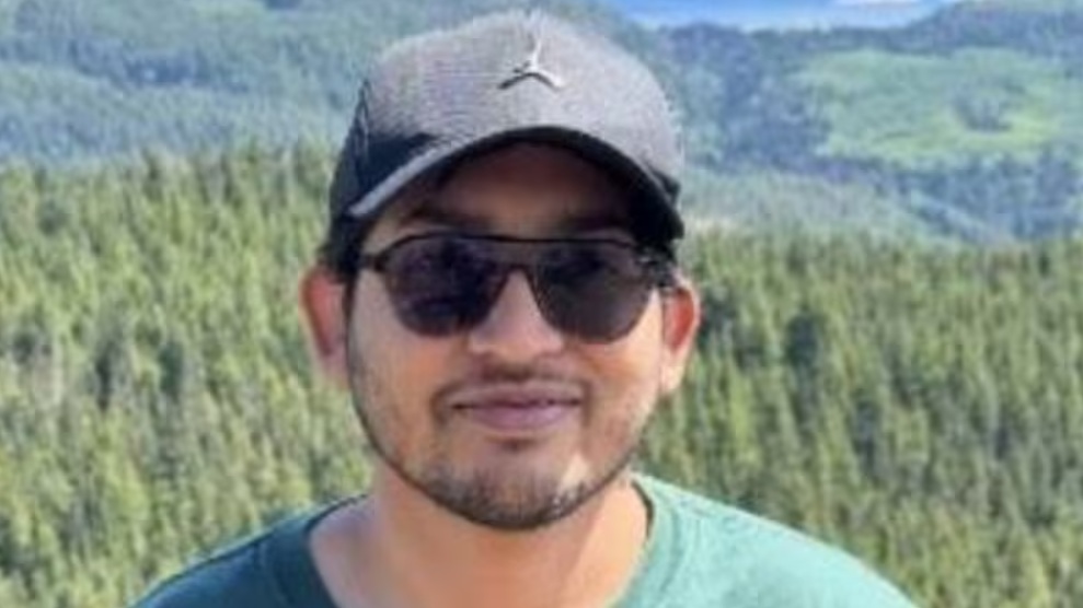 Man wearing a dark cap and sunglasses outdoors with a forested mountainside behind him