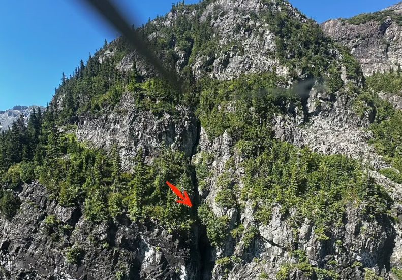 Steep rocky mountainside with evergreen trees and a red arrow pointing to a narrow crevice or cave opening in the rock face.