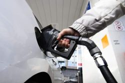 Person fueling a white car at a gas station, nozzle inserted into the fuel port.