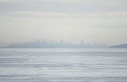 Hazy city skyline across calm water on a misty day, with indistinct high-rise buildings along the horizon.