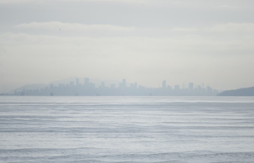 качество воздуха улучшится | Наш Ванкувер Hazy city skyline across calm water on a misty day, with indistinct high-rise buildings along the horizon.