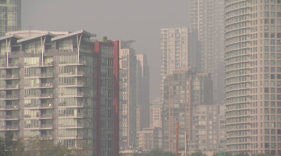 Hazy city skyline with modern high-rise apartment buildings lining the street. A dense row of glass and concrete towers viewed through smog or haze, typical of an urban residential district.