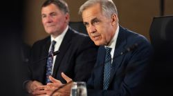 Two men in suits sit at a panel table; one speaks into a microphone while the other listens.