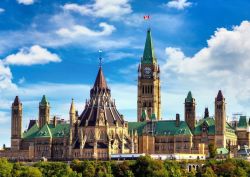 Parliament of Canada buildings with green roofs and the tall Peace Tower against a blue sky, flag waving atop.
