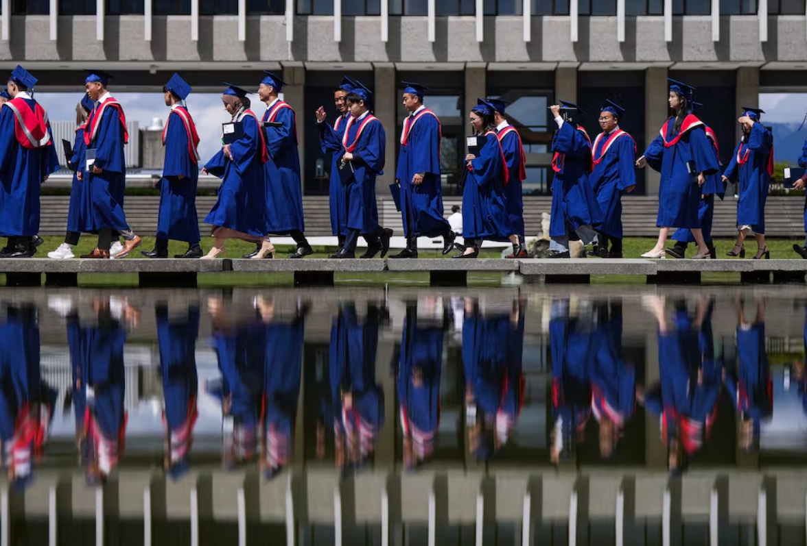 Graduates in blue gowns with red sashes walk in a line beside a reflecting pool, buildings in the background.