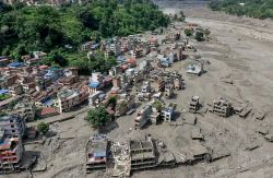 A densely built hillside town on a muddy riverbank with many damaged and collapsed buildings amid debris after a disaster.