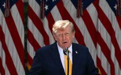 Man in a navy suit and yellow tie speaks at a lectern with multiple American flags in the background, likely at a formal event.