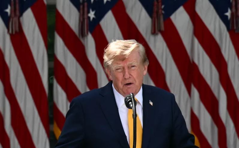 Man in a navy suit and yellow tie speaks at a lectern with multiple American flags in the background, likely at a formal event.