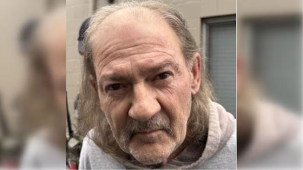 Close-up of an older man with long gray hair and a mustache, wearing a light gray hoodie outdoors near a building.