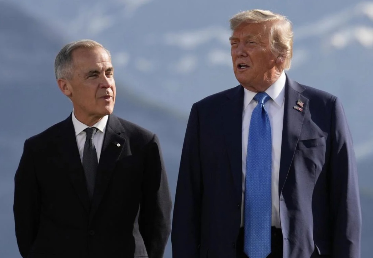 Two men in dark suits stand outdoors, mountains in the background, engaged in conversation.