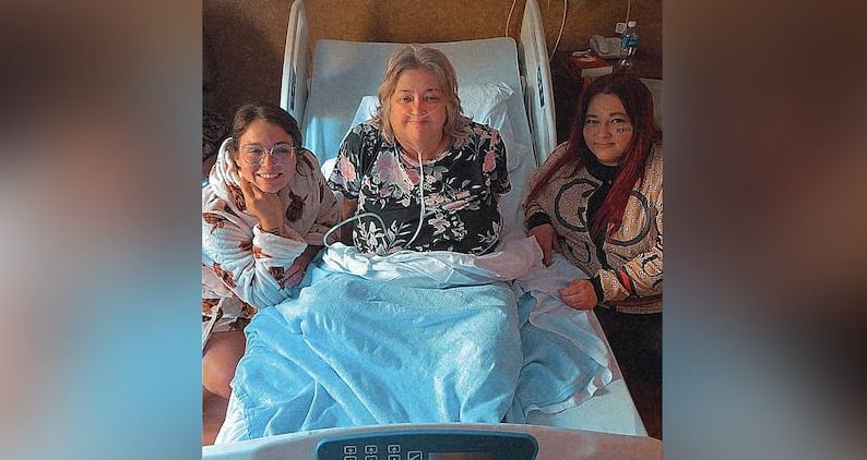 Three women sit beside a hospital bed: the patient with an oxygen tube and a blue blanket, flanked by two visitors smiling.