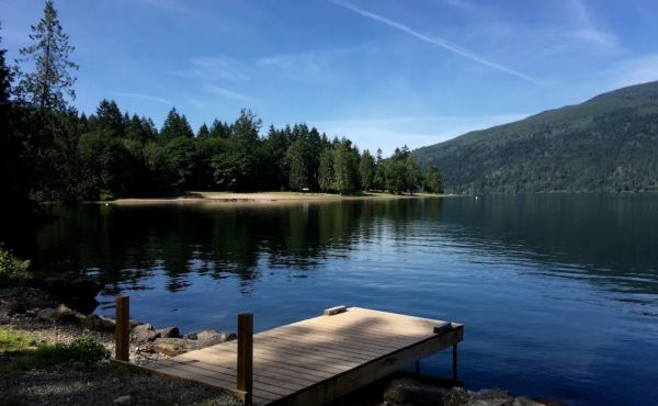 A wooden dock extends into a calm lake, with a forested shore and mountains under a clear blue sky.