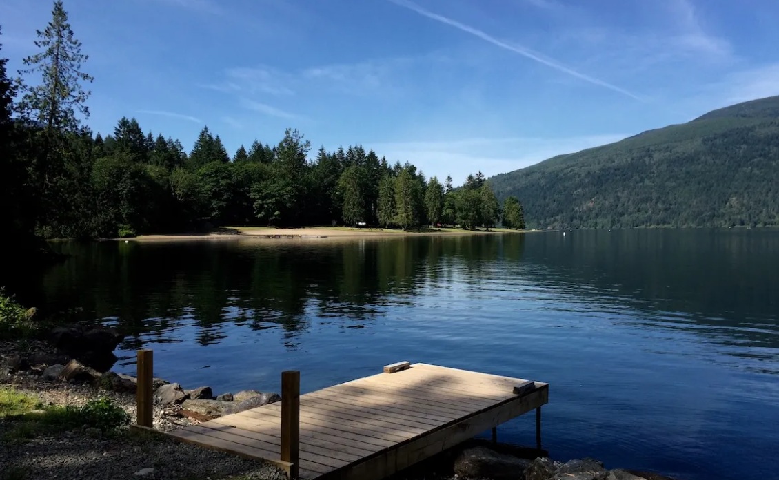 A wooden dock extends into a calm lake, with a forested shore and mountains under a clear blue sky.