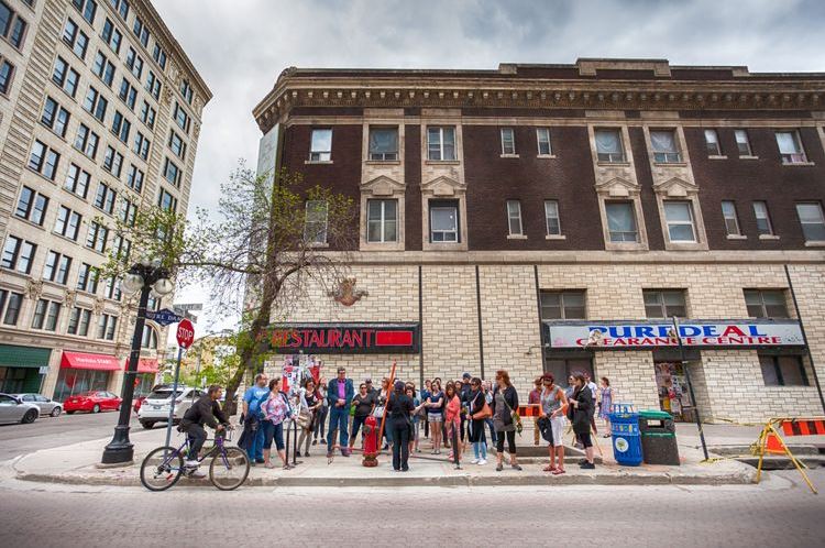 A group of people gathered on the sidewalk in front of a brick building with a restaurant sign, as a cyclist rides past on the street to the left.