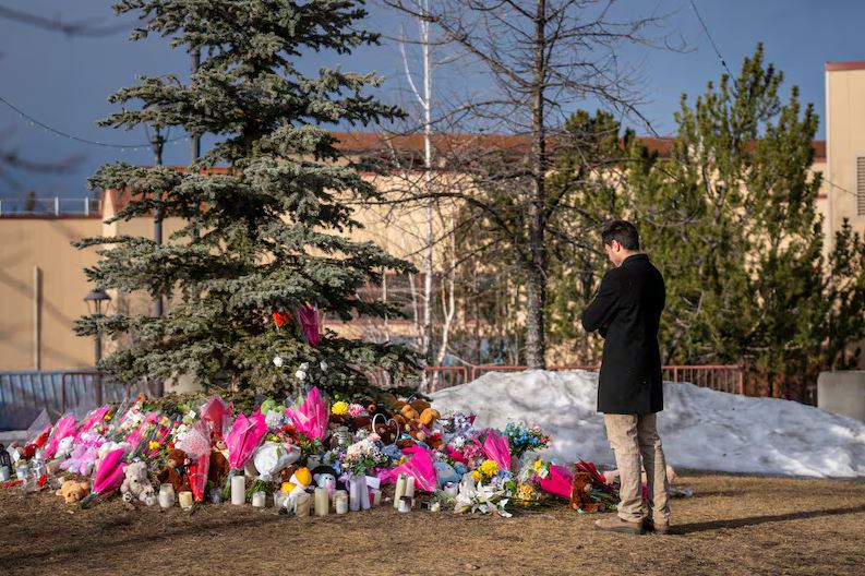 Мемориал посвященный жертвам стрельбы в средней школе Тамблер-Ридж | Наш Ванкувер Man in a dark coat stands with his back to the camera at a memorial site with flowers and candles around a tree.