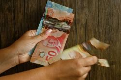 Two hands count a fan of Canadian banknotes on a dark wooden surface (includes 5, 20, 50 dollar bills).
