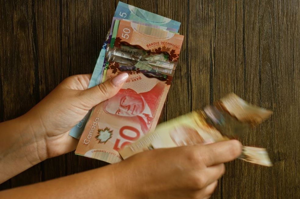 Two hands count a fan of Canadian banknotes on a dark wooden surface (includes 5, 20, 50 dollar bills).