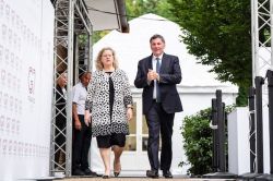 Two well-dressed individuals walk off a stage at an outdoor event, a woman in a patterned coat and a man in a dark suit.