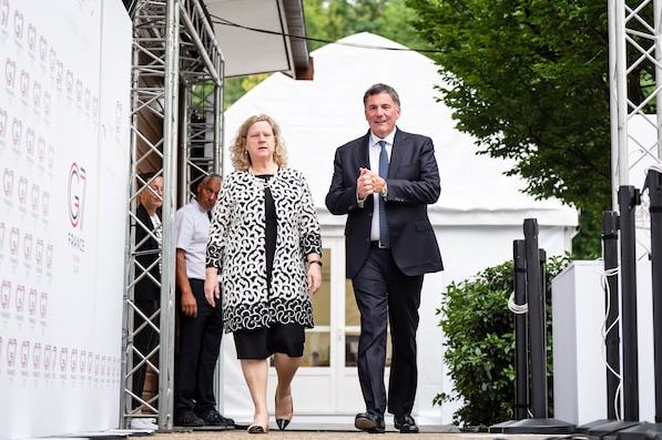 Two well-dressed individuals walk off a stage at an outdoor event, a woman in a patterned coat and a man in a dark suit.