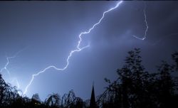 Bright jagged lightning bolt across a dark night sky above silhouetted trees and a church steeple.