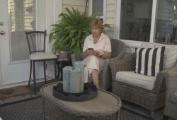 Elderly woman on a wicker porch chair, looking at her smartphone, with a fern behind and candles on the table in front of her.