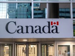 Sign reading 'Canada' with a small Canadian flag above, at the entrance of a modern glass-fronted building.