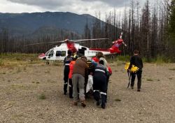 Emergency responders wheel a stretcher toward a red-and-white rescue helicopter on a rocky airstrip with a burned forest in the background and mountains beyond.