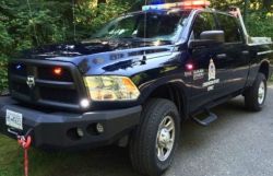 Dark blue police pickup truck with red and blue emergency lights on the roof, parked on a wooded roadside near the front bumper with a winch rope.
