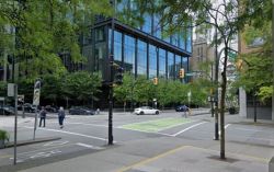 Street intersection in a city with a glass-fronted office building, green trees, and pedestrians crossing.