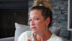 Woman with a messy bun wearing a white cardigan sits on a sofa, looking thoughtful with a hand near her chin.