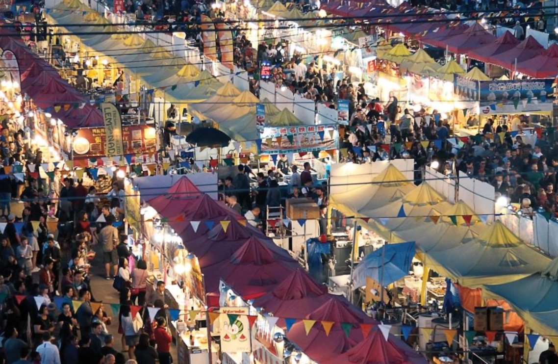 Aerial view of a crowded outdoor night market with colorful tents and string lights.