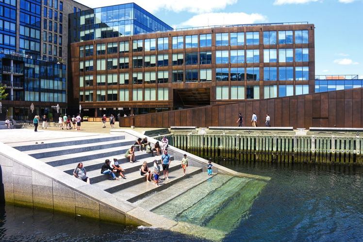 Modern glass-and-brick office building along a waterfront with tiered concrete steps where people sit and relax.