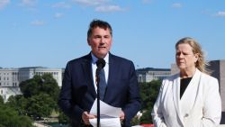 Man in a navy suit speaks at a microphone with papers in hand, flanked by a woman in a white blazer outdoors with government buildings in the background.
