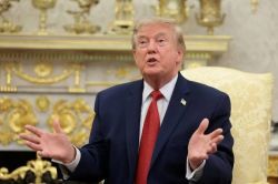 Man in a dark suit and red tie sits in an ornate, gold-decorated room, gesturing as he speaks.