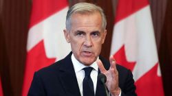 Man in a dark suit speaking at a podium with a microphone, Canadian flags in the background.