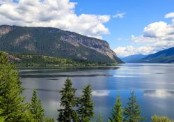 Wide lake surrounded by forested mountains under a blue sky with fluffy clouds, daytime scene.