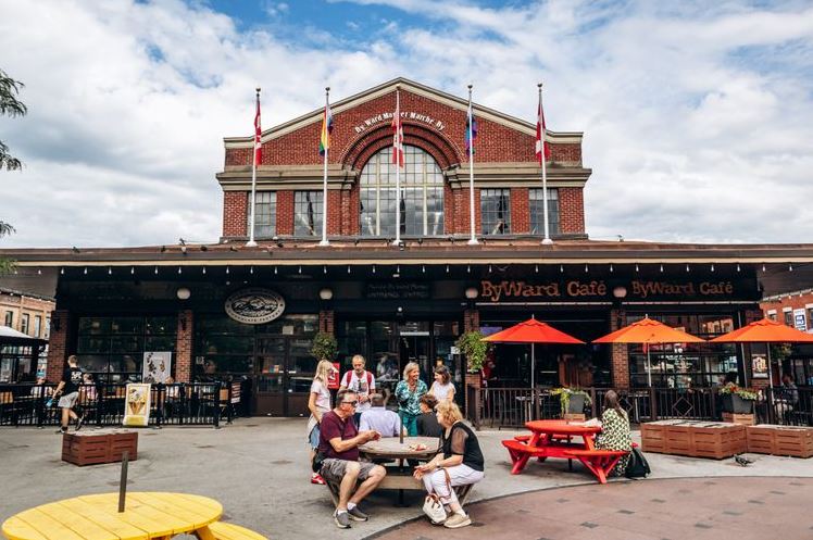 Brick ByWard Café building with outdoor seating, red umbrellas, and people gathered around a table in a plaza.**Note: remove the stray asterisks in final output**