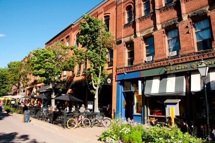 Brick storefronts line a sunny street with outdoor cafe seating under umbrellas and bicycles parked nearby.
