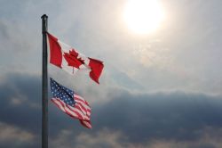 Canadian flag and American flag fluttering on a flagpole against a cloudy sky and bright sun.
