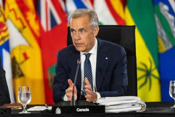Man in a navy suit speaks into a microphone at a formal briefing, with colorful flags behind and a stack of papers on the desk.