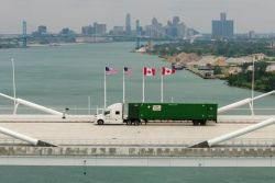 White semi-truck pulling a green shipping container crosses a bridge over a river, flagpoles with US and Canadian flags line the road, distant city skyline visible.