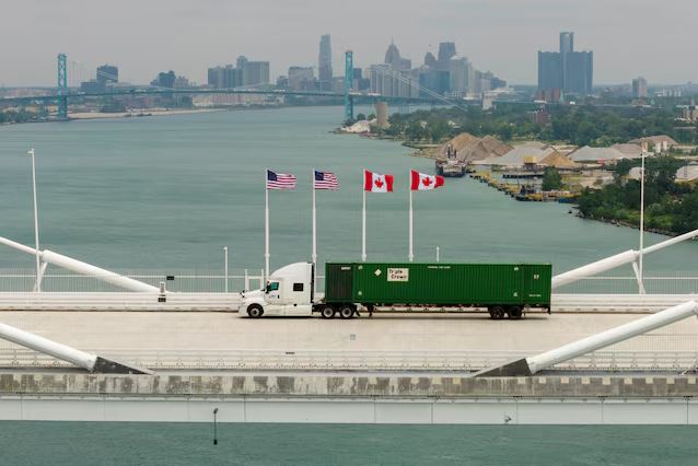 Ответные тарифы | Наш Ванкувер White semi-truck pulling a green shipping container crosses a bridge over a river, flagpoles with US and Canadian flags line the road, distant city skyline visible.