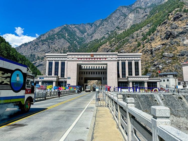 Grand gateway and government building at a mountain border area, with a wide road, vehicles, and colorful flags in front; rugged mountains rise behind the structure.