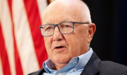 Close-up of an older man wearing blue glasses and a light blue shirt under a dark blazer, speaking, with a blurred red-and-white flag in the background.