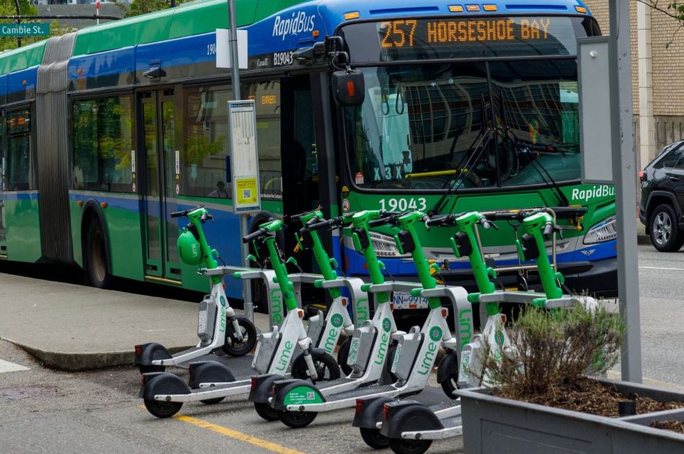 City bus at a stop with a row of lime-colored Lime electric scooters parked in front on a curbside downtown street.