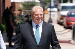 Smiling older man in a dark suit and blue tie walks outdoors on a sunny city street.