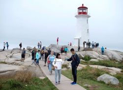 White lighthouse with red top on a rocky coastline, with groups of tourists walking along a path on a misty day.
