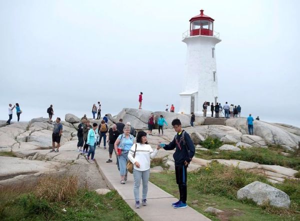 White lighthouse with red top on a rocky coastline, with groups of tourists walking along a path on a misty day.