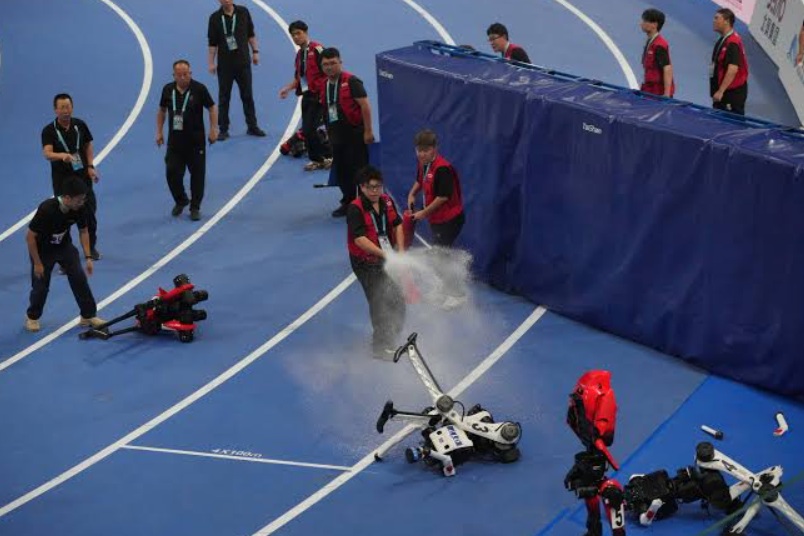 Team members in red vests push a small robot on a blue competition track while smoke rises from the crash site and another robot lies on the floor nearby.